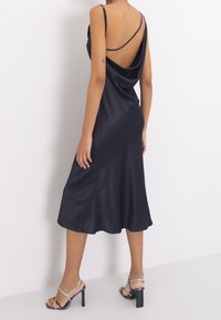 Black satin slip dress with a draped back and thin shoulder straps. Features a mid-length cut and a smooth texture. Paired with multicolored heels.