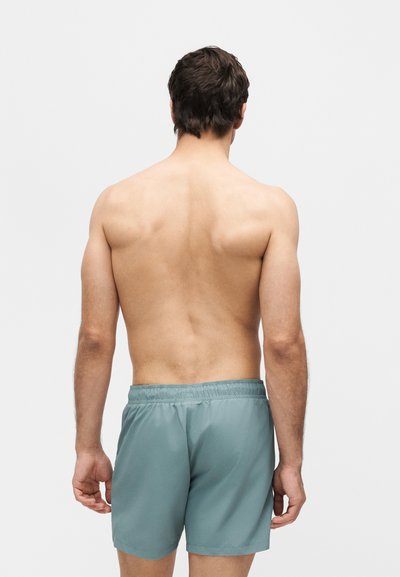 Άνδρας που στέκεται με την πλάτη, χωρίς πουκάμισο, φορώντας ανοιχτό μπλε μαγιό, με απλό λευκό φόντο.