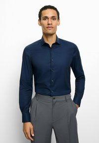 Dunkelblaues Hemd mit Kragen, langen Ärmeln und Knopfleiste, kombiniert mit grauen Hosen. Glatter Stoff mit dezenter Struktur.