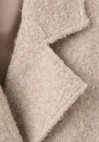 Abrigo de lana de color beige claro con una superficie texturada y suave; presenta un solapa afilada y bordes sin rematar. Enfóquese en el material suave y cálido.