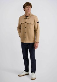 Beige knoopjack met twee borstzakken, zwarte knopen en een gestructureerde afwerking. Gekleed met marineblauwe broek en witte sneakers.