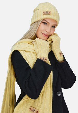 Berretto giallo lavorato a maglia, guanti senza dita e sciarpa con texture a treccia, con un'etichetta in similpelle. Indossato con un cappotto nero.