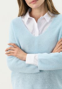 Lichtblauwe gebreide trui met een V-hals en lange mouwen, gedragen over een witte blouse met kraag. Heeft een gestructureerde, losse weving.