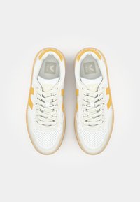 Sneakers bianchi con dettagli gialli, texture perforata e suole in gomma beige. Presentano un logo grigio sulla linguetta e chiusura con lacci.