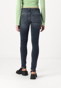 Sötét farmer skinny farmere, magas derékkal, hátulról bemutatva, fekete magassarkú szandálokkal kombinálva. Zöld, bordázott felső látható.