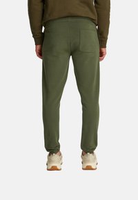 Pantalones de chándal verde oliva hechos de una tela suave, con piernas ajustadas, cintura elástica y un bolsillo trasero, combinados con zapatillas beige.