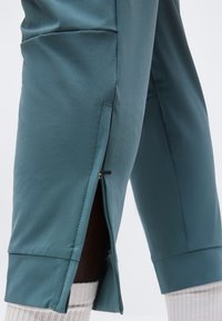 Κοντινό πλάνο στο πόδι από τυρκουάζ παντελόνι με πλευρικό φερμουάρ που είναι μερικώς ανοιχτό, αποκαλύπτοντας μαύρο ύφασμα από κάτω και λευκές ριμπ πλεκτές κάλτσες.