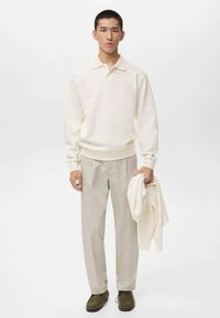 Cremfarbener Polo-Pullover mit lockerer Passform, beiger maßgeschneiderter Hose, olivgrüne Schuhe. Das Modell hält einen leichten Pullover und steht vor einem neutralen Hintergrund.