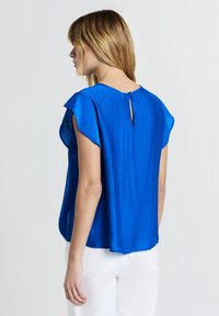 Donna con capelli biondi fino alle spalle, che indossa una blusa blu reale a maniche corte e pantaloni bianchi, vista da dietro su uno sfondo bianco.
