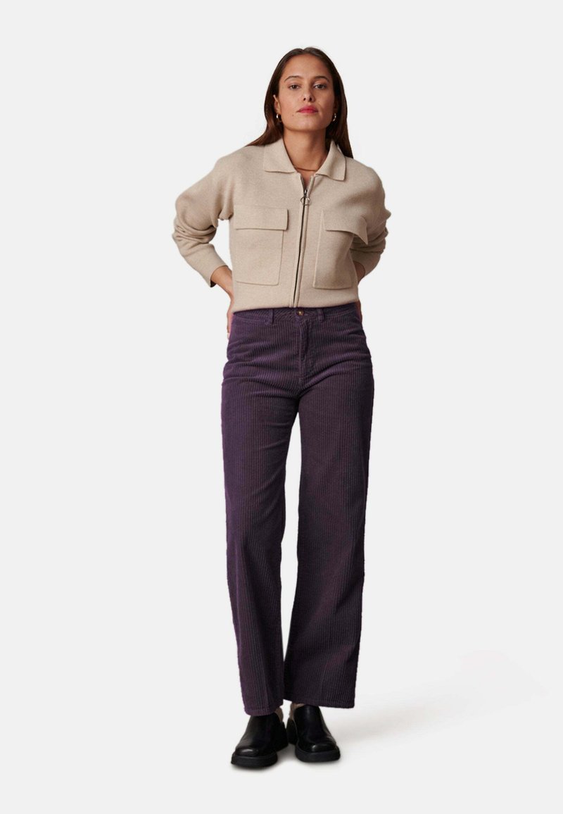 Beige ritsvest met twee voorzakken, gecombineerd met paarse flanellen wijde broek. Model staat in zwarte enkellaarsjes.
