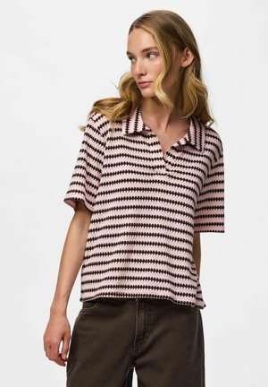 Jeune femme aux longs cheveux blonds portant une chemise à manches courtes rayée rose et noire avec col et un pantalon foncé, regardant vers sa gauche.