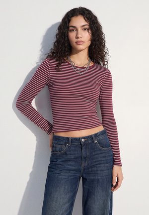 Mladá žena s kučeravými vlasmi nosí dlhý rukáv bordovo-biele pruhované crop top a tmavomodré džínsy, stojí pri bielej stene.