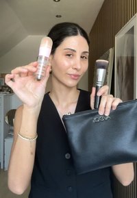 Fond de teint avec un capuchon rose, un pinceau à estomper et une trousse en cuir noir avec le logo "ZOOVA". Textures lisses et tons neutres.