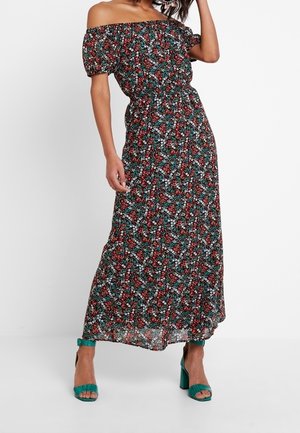 Kobieta ubrana w długą sukienkę w kwiaty z odkrytymi ramionami i bufiastymi rękawami oraz turkusowe sandały na słupku z odkrytymi palcami.