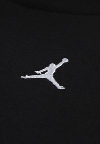 Zwarte stof met een wit geborduurd Jumpman-logo, waarop een figuur midden in de lucht met een basketbal wordt afgebeeld, met fijne details en textuur.