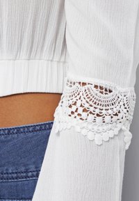 Blusa bianca, strutturata, con maniche lunghe e dettagli in pizzo intrecciato sui polsini. Un paio di jeans in denim blu scuro è parzialmente visibile.