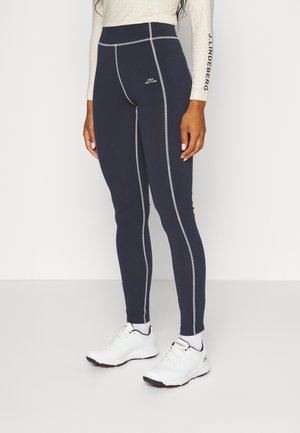 Frau trägt schwarze Leggings mit kontrastierenden Nähten, weiße Sportschuhe und ein gemustertes Langarmshirt mit Logo auf dem Ärmel.