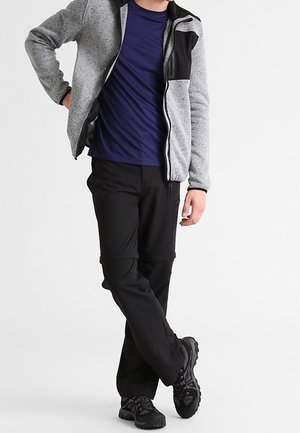 Persona che indossa una giacca grigia con cerniera sopra una maglietta blu scuro, pantaloni neri con zip e scarpe da trekking nere, in piedi con le gambe incrociate e una mano sul fianco.