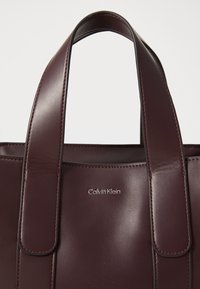 Burgundi odos rankinė su dviem rankenomis, lygaus paviršiaus, su iškilniu "Calvin Klein" logotipu priekyje. Paprastas dizainas be puošmenų.
