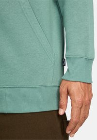 Felpa verde chiaro con polsini e orlo a coste, dotata di una tasca laterale e dettaglio di tessuto testurizzato. Mano visibile su uno sfondo marrone.