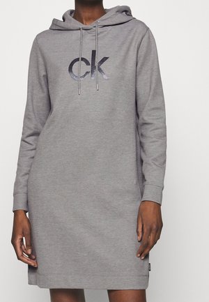 Personne portant une robe grise à manches longues avec capuche, imprimée du logo "ck" sur la poitrine et munie de cordons de serrage à la capuche.
