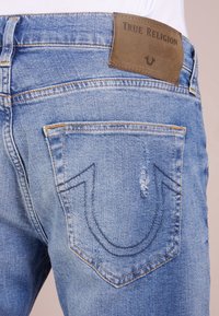 Ljusblå denimjeans med en läderpatch från True Religion, kontrasterande sömmar och en signaturdesign av hängslet på bakfickan.