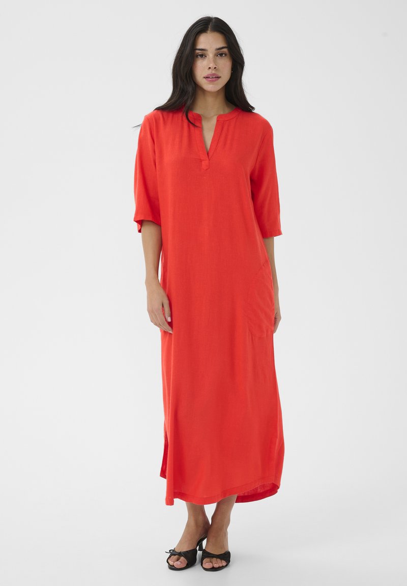 Frau steht und trägt ein lockeres, knielanges, leuchtend rotes Kleid mit dreiviertel Ärmeln und schwarzen offenen Pumps vor einem schlichten weißen Hintergrund.