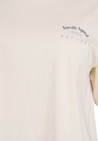 Beige bomulds t-shirt med rund halsudskæring, korte ærmer og et trykt marineblåt design, der inkluderer "Varsity Squad SPORTS EST. 1980."
