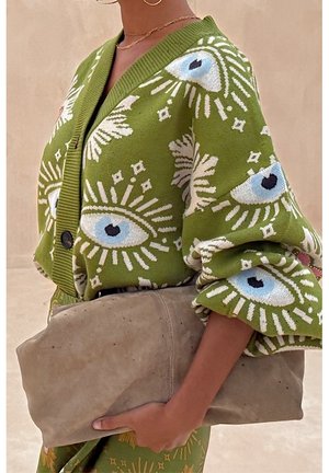 Personne portant un cardigan vert avec des motifs d'yeux blancs et bleus, tenant une grande pochette beige en daim sur fond neutre.