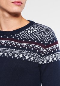 Marineblauwe gebreide trui met een ronde halslijn, voorzien van witte en bordeauxrode geometrische patronen over de schouders en de bovenborst.