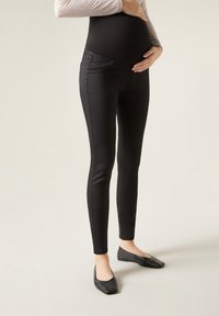 Svarta gravidleggings med en slät yta, hög midja och framfickor, som bärs med platta svarta skor mot en neutral bakgrund.