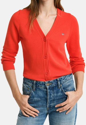 Kvinde iført en lyserød rød knappet cardigan med ribstrikkede manchetter og højtaljede blå jeans, hænder hvilende nær lommerne.