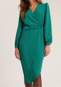 Femme portant une robe bleu sarcelle à manches longues, longueur genou, avec un devant croisé et un détail froncé à la taille, debout devant un fond uni.