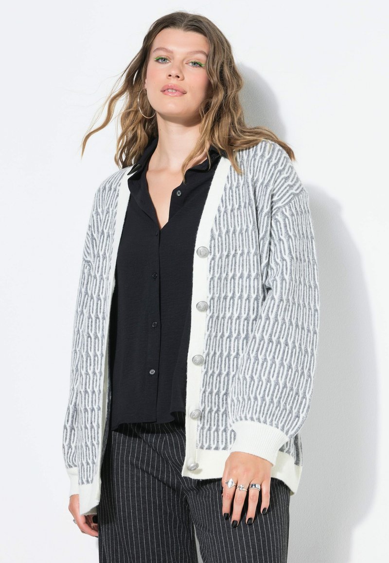 Cardigan grigio e bianco a motivo con chiusura a bottone, dotato di polsini a coste e una vestibilità rilassata, indossato sopra una maglietta nera e pantaloni a righe.