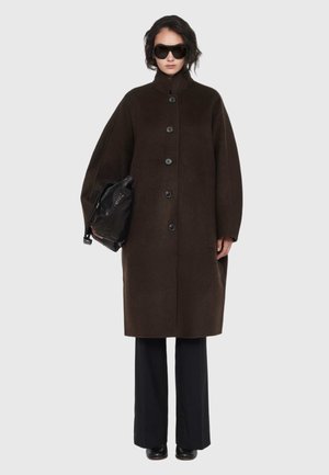 Vrouw draagt een oversized donkerbruine jas met knopen, zwarte wijde broek, grote zonnebril en houdt een zwarte leren tas vast tegen een effen achtergrond.