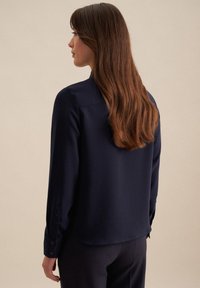 Navyblaue Bluse mit langen Ärmeln, glatter Textur, Knopfmanschetten und abgerundetem Saum, von hinten gesehen.