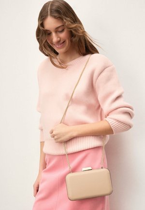 Joven mujer que lleva un suéter y falda rosa suave, sosteniendo un bolso rectangular beige con una correa de cadena dorada, sonriendo y mirando hacia abajo.