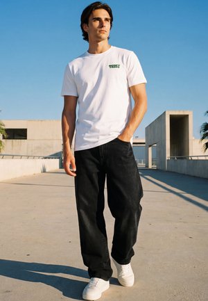 Junger Mann trägt ein weißes "Money Trees" T-Shirt, schwarze Hose und weiße Sneakers, steht an einem sonnigen Tag draußen vor modernen Betongebäuden.