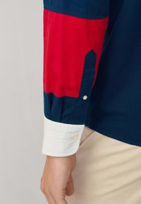 Camisa de manga larga multicolor con secciones en azul marino, rojo y blanco, con un puño de botón blanco y diseño de manga estructurada.