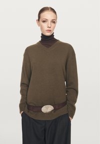 Donna con un maglione a V verde oliva sopra un dolcevita marrone scuro, pantaloni larghi scuri, mani nelle tasche, con una cintura caratterizzata da una grande fibbia ovale.