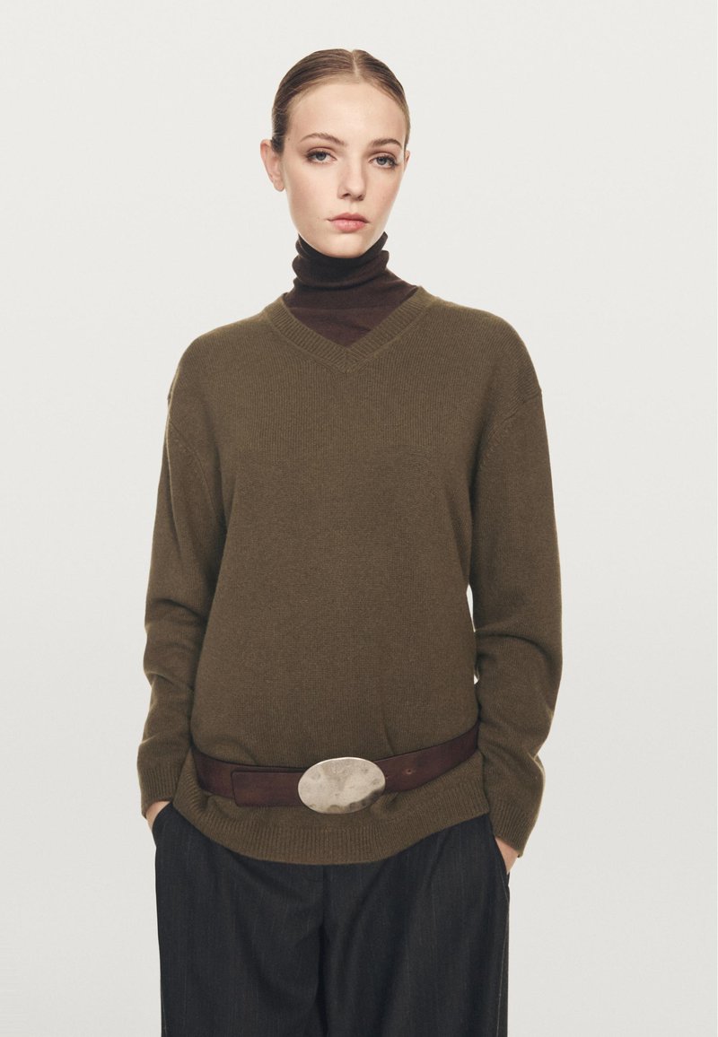 Donna con un maglione a V verde oliva sopra un dolcevita marrone scuro, pantaloni larghi scuri, mani nelle tasche, con una cintura caratterizzata da una grande fibbia ovale.