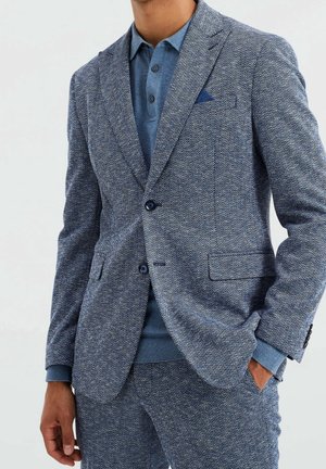 Blazer a motivi a spina di pesce blu abbinato a un polo azzurro chiaro e pantaloni strutturati coordinati. Presenta rever a lancia e due bottoni.