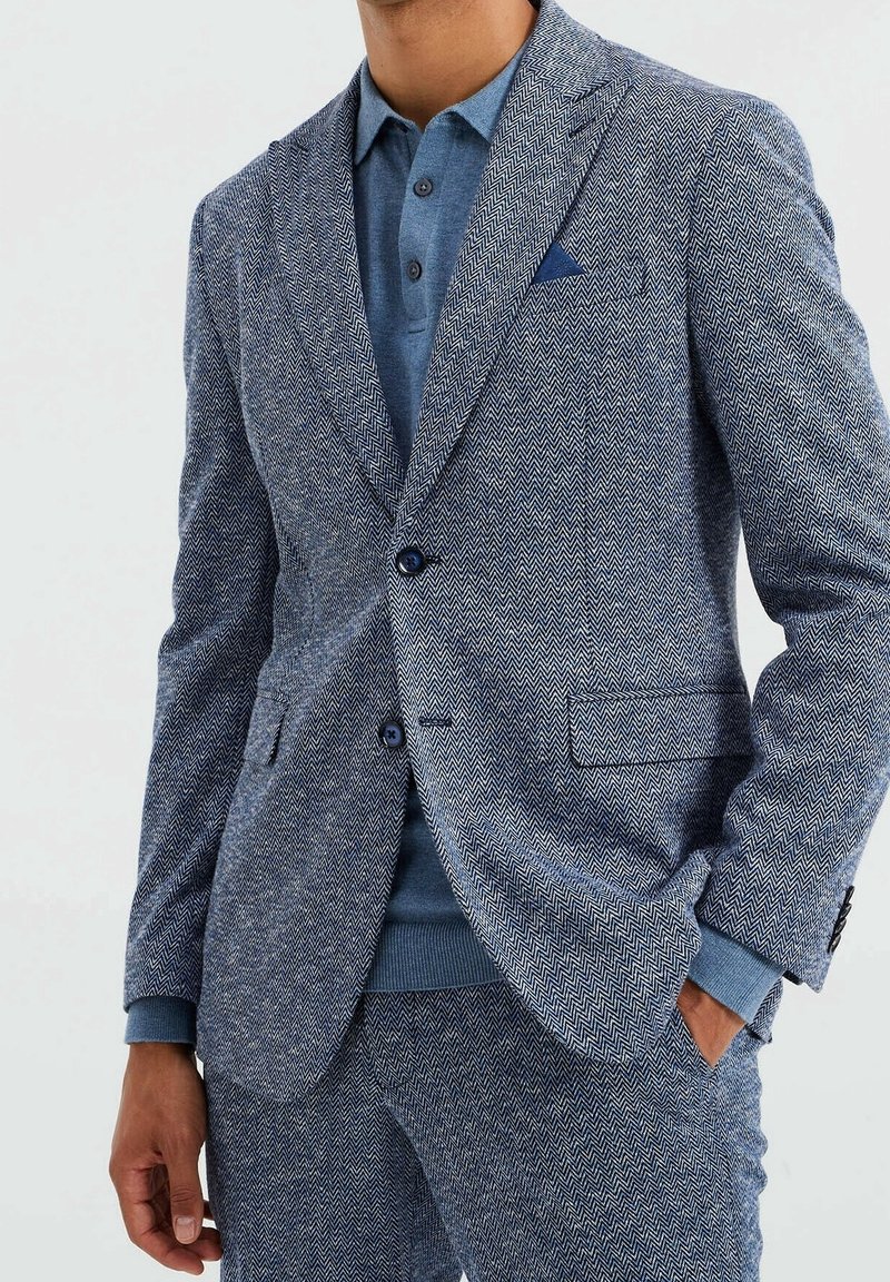 Blå blaser med fiskbensmönster, parat med en ljusblå polo och matchande texturerade byxor. Har slagna lapeller och två knappar.