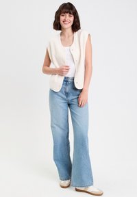 Gilet sans manches en crème et tout en douceur avec des boutons-pression ; associé à un jean évasé bleu clair et des baskets beiges. Design simple et décontracté.
