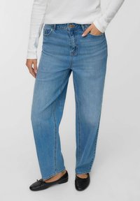 Donna che indossa jeans dritti azzurri chiari con una maglietta bianca a maniche lunghe infilata dentro e ballerine nere, in piedi con una mano in tasca.