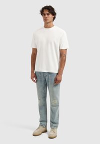 Man staat in een eenvoudige witte T-shirt met korte mouwen, lichte blauwe distressed jeans en beige veterschoenen, tegen een lichtgrijze achtergrond.