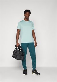 Camiseta deportiva azul claro de manga corta, combinada con pantalones de chándal verde oscuro y zapatillas negras, sosteniendo una mochila de gimnasio negra.
