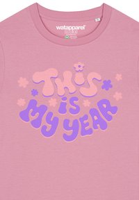 Lichtroze katoenen t-shirt met vetgedrukte, kleurrijke tekst: "Dit is Mijn Jaar" in paarse en perzikachtige bubbelfonten, verfraaid met kleine bloemetjes graphics.
