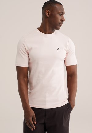 Férfi világos rózsaszín rövid ujjú pólóban, kis fekete madár logóval, sötét nadrágban, jobbra néz, egyszínű bézs háttér előtt.