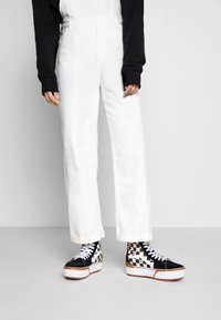 Pantalones de mezclilla blancos con corte recto, combinados con zapatillas altas de cuadros en blanco y negro con cordones blancos y suela gruesa.